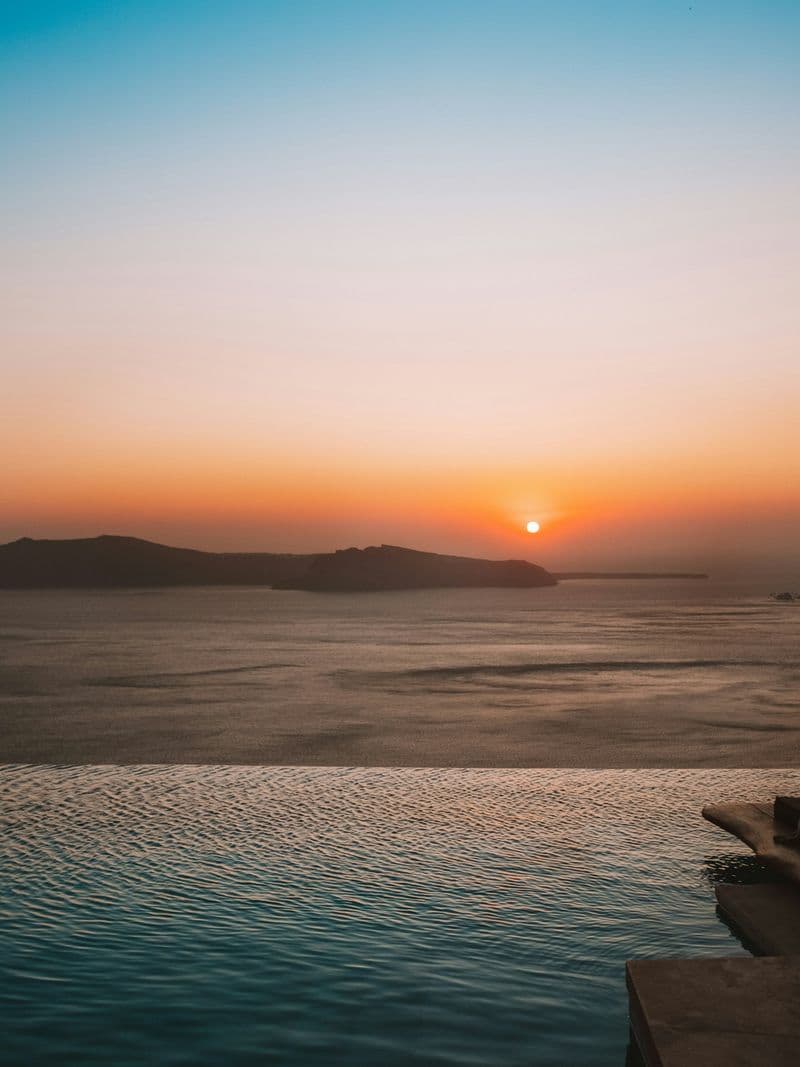 Infinity pool overlooking the ocean at golden hour, with the sun setting behind a distant island silhouette