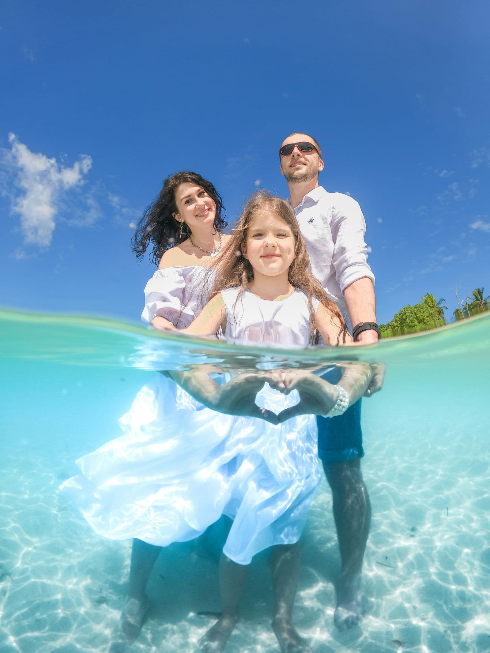 Split-level underwater/above-water shot of a family standing in clear turquoise tropical sea — parents and a young daughter, white sky and palm-trimmed shore behind them