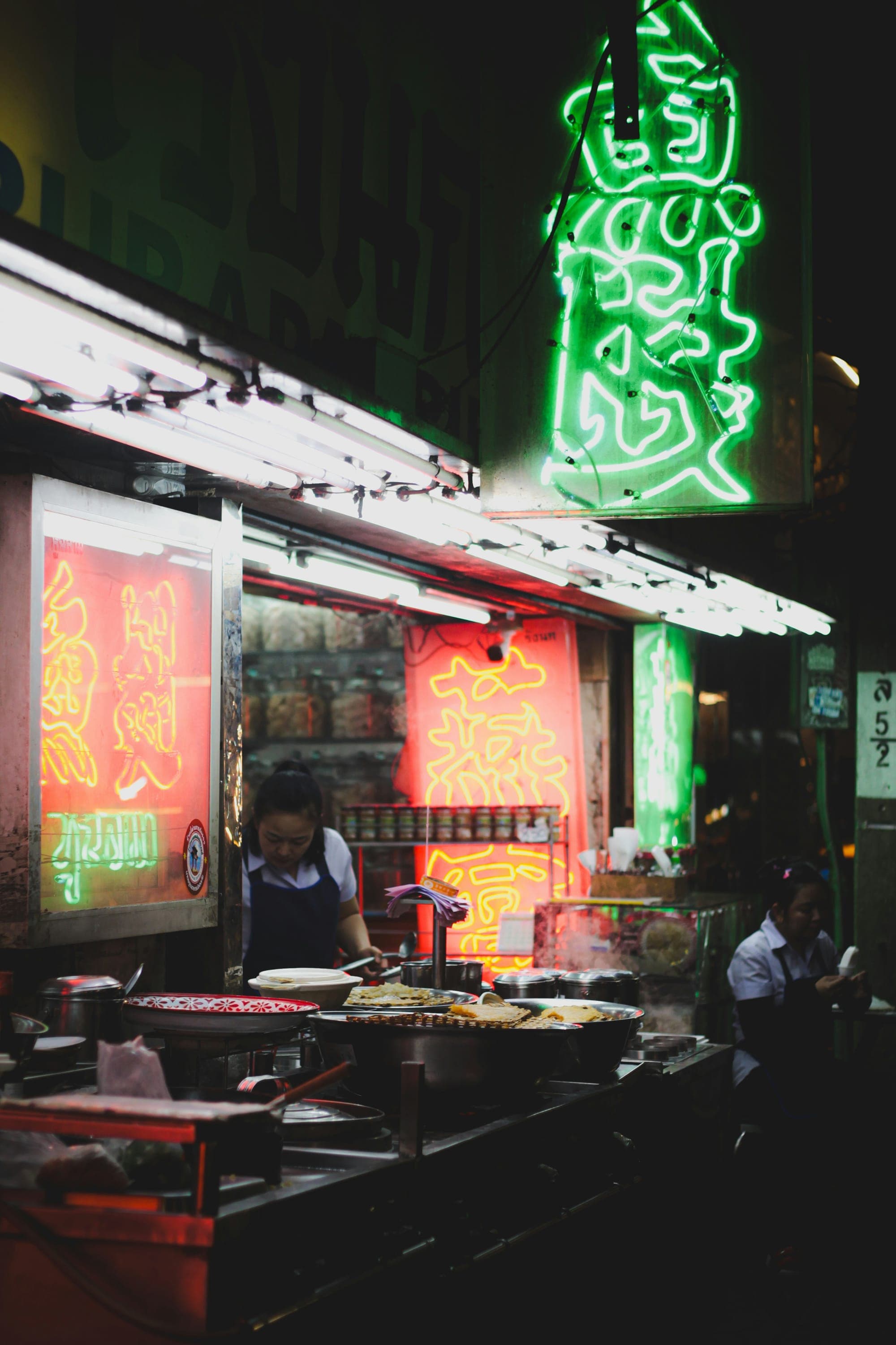 Southeast Asian night market food stall with glowing neon signs and street vendors in the evening
