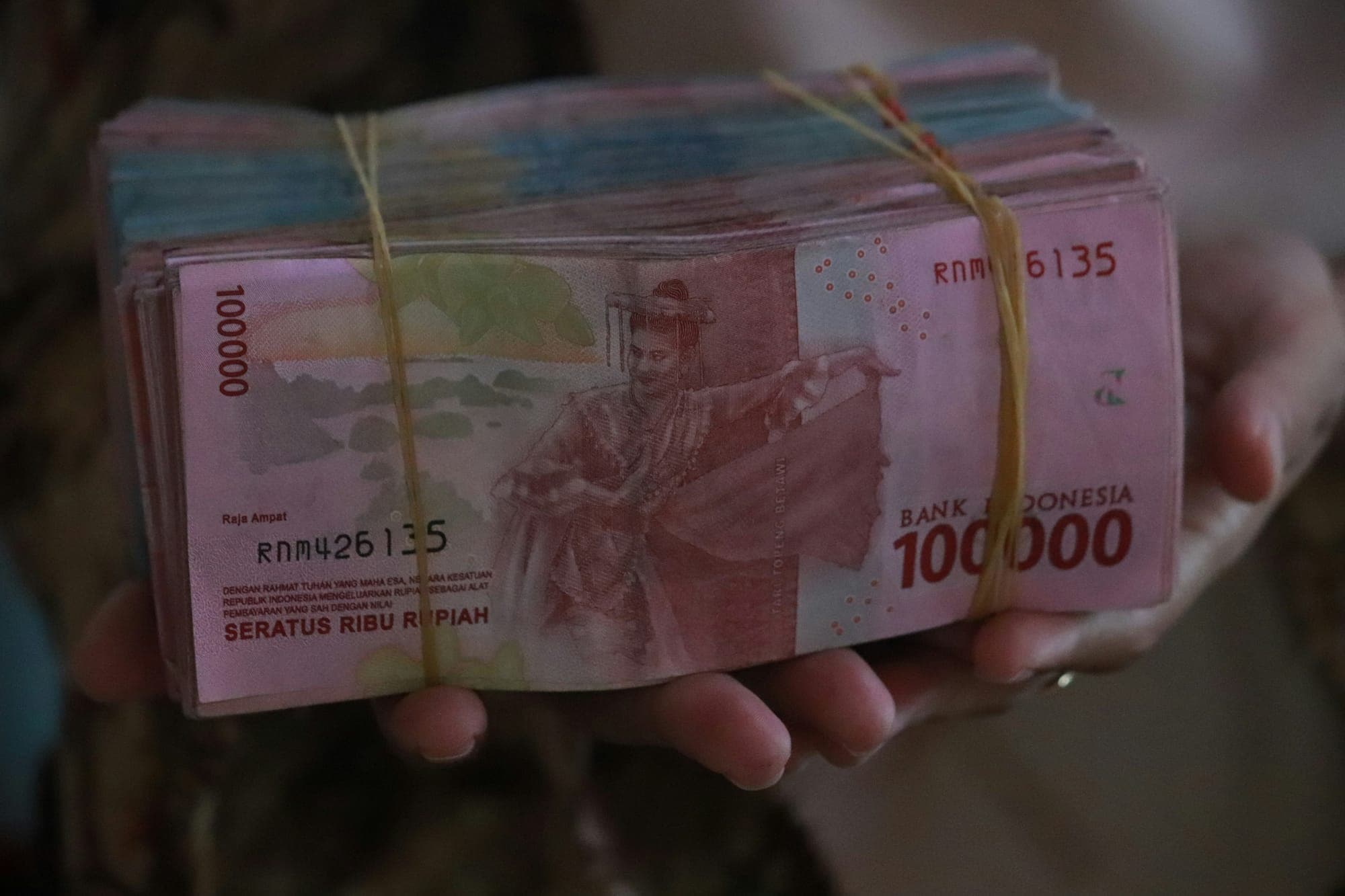 A hand holding a stack of Indonesian Rupiah 100,000 banknotes bundled with a rubber band — the highest-denomination currency note used in Batam